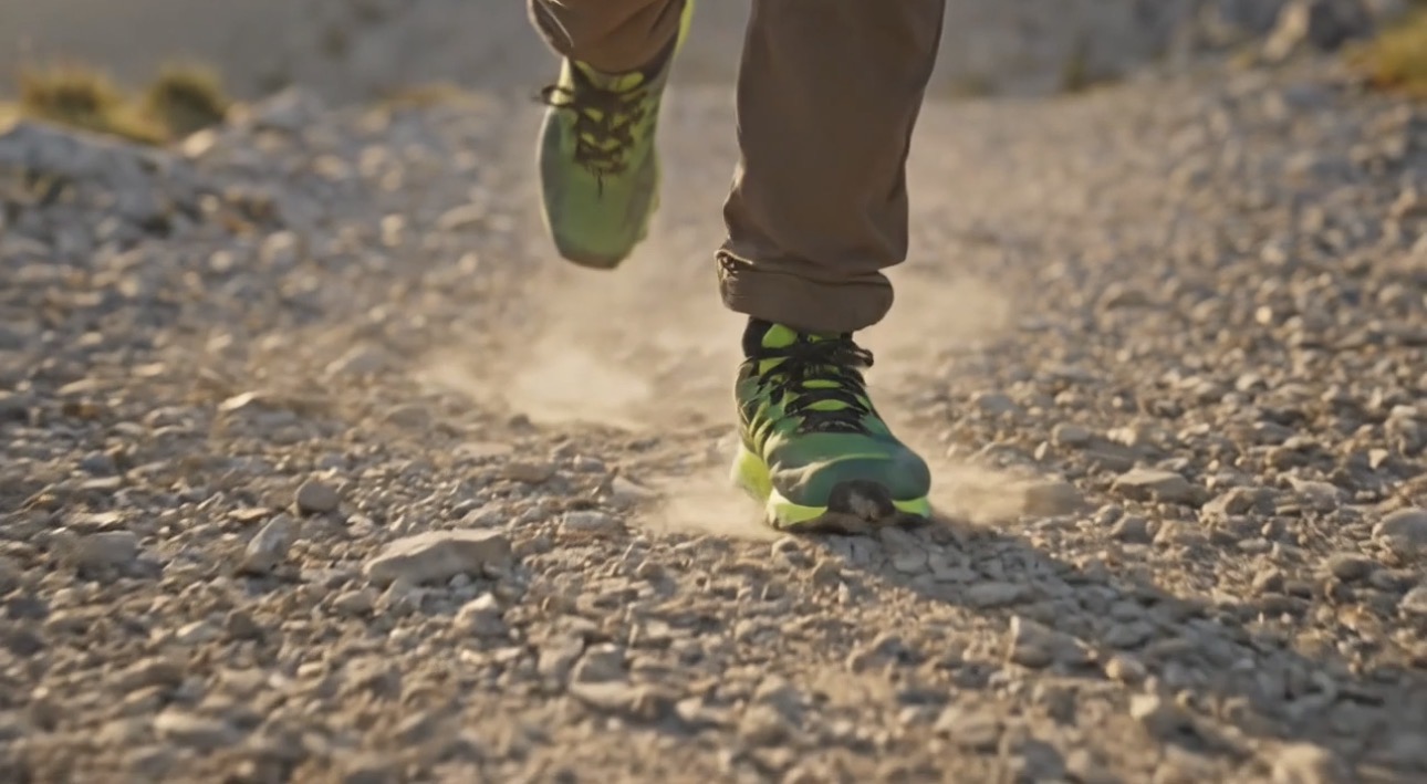 Video still: low-angle close-up of neon green trail shoes kicking up dust on rocky path